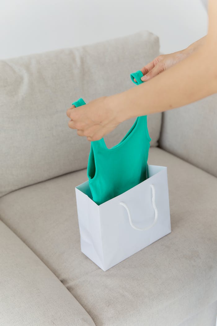 Person unpacking a green tank top from a white shopping bag on a beige couch.